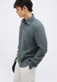 Νέος άνδρας με σκούρα σγουρά μαλλιά, φοράει πουκάμισο σε σκούρο πράσινο χρώμα με κουμπιά και ανοιχτό μπεζ παντελόνι, κρατώντας μια μικρή μαύρη τσάντα, κοιτάζοντας προς τα αριστερά.