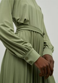 Långärmad, ljusgrön klänning av mjukt tyg med skärp i midjan, plisserad kjol och knäppta ärmslut. Mjuk textur, elegant design.