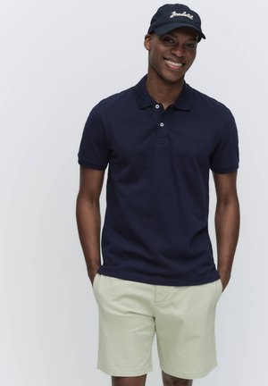 Uśmiechnięty mężczyzna w granatowej koszulce polo, beżowych szortach i czarnej czapce z daszkiem, stojący z rękami w kieszeniach na białym tle.