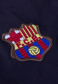 FC Barcelona voetbalclub geborduurd embleem met rode en blauwe strepen, een gele bal en een rood kruis op een donkere stofachtergrond.
