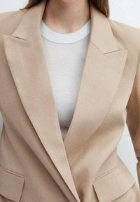 Beige lněný blazer s ostrými klopami a přizpůsobeným střihem, nošený přes světle šedé žebrované tričko. Obsahuje kapsy a texturovanou látku.