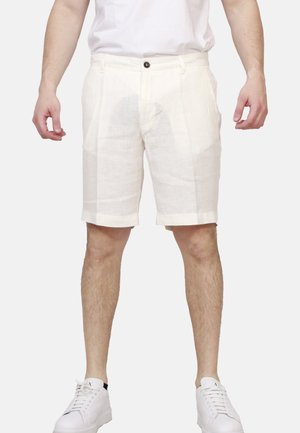 Uomo con pantaloncini bianchi fino al ginocchio, camicia bianca e scarpe da ginnastica bianche, in piedi davanti a uno sfondo neutro.