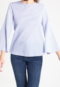 Top a righe blu chiaro e bianche, a maniche lunghe con scollatura rotonda e maniche a campana; aderente sul busto e morbido sotto.