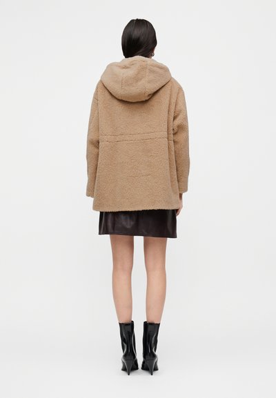 Καφέ ζακέτα με επένδυση shearling και κουκούλα, με άνετη εφαρμογή, χωρίς ραφές και υφή ύφασμα. Φορεμένη πάνω από μια σκουρόχρωμη δερμάτινη φούστα.