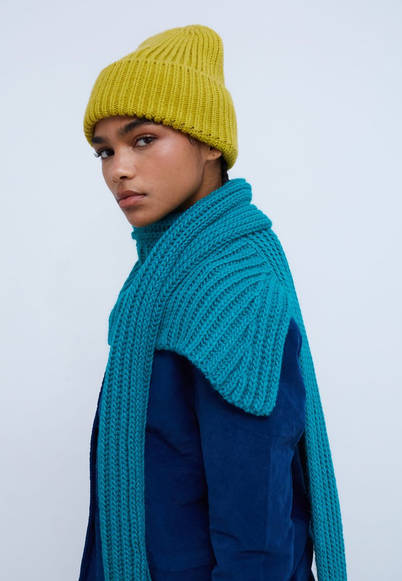Tyrkysová pletená šál s rebrovanou textúrou, doplnená žltou rebrovanou čiapkou, vrstvená cez tmavomodrú bundu.