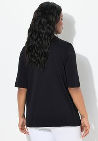 Top nero a maniche corte realizzato in tessuto liscio. Presenta una vestibilità ampia con fessure laterali e un collo tondo, mostrato da dietro.