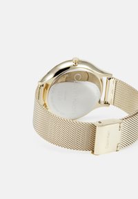 Montre en acier inoxydable doré avec un boîtier arrondi, un fond transparent et une fermeture. Présente un design minimaliste et une texture lisse.