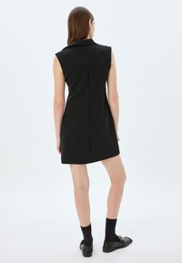 Zwarte mouwloze jurk met een kraag, een aansluitende silhoeët en details in de achternaad, gecombineerd met zwarte sokken en schoenen. Gladde textuur.