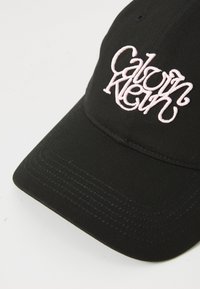 Zwarte katoenen baseballpet met een gebogen rand en een roze geborduurd "Calvin Klein" logo aan de voorkant. Geweven stof met stikseldetails.