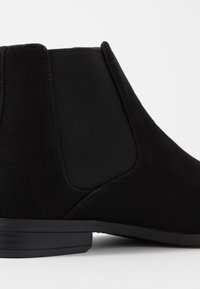 Bota de tornozelo em camurça preta com painéis laterais elásticos, sola plana preta e um perfil limpo. Apresenta detalhes de costura subtis.