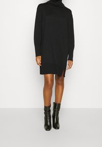 Schwarzes Rollkragenstrickkleid mit gerippten Bündchen und Saum, ausgestattet mit seitlichen Schlitzen. Tragen mit schwarzen Knöchelstiefeln.