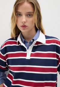 Joven mujer con cabello rubio, vistiendo una camiseta de rugby a rayas rojas, azules y blancas sobre una camisa de cuadros azul.