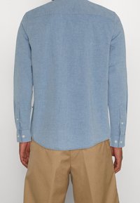 Chemise à manches longues bleu clair en tissu doux, avec un ourlet courbé, des poignets à boutons et un design de jaser arrière simple.