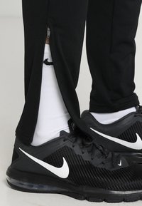 Schwarze Sporthose mit Reißverschlüssen an den Knöcheln, kombiniert mit schwarzen Turnschuhen mit einem weißen Nike-Swoosh und weißen Socken mit einem Logo.