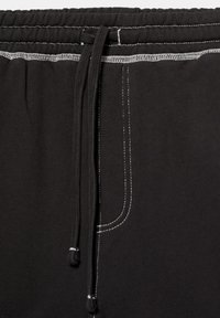 Gros plan de la ceinture noire d’un pantalon de survêtement avec des cordons noirs et des surpiqûres blanches visibles le long de la taille et des bords des poches.
