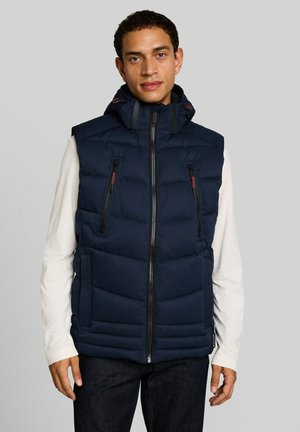 Uomo che indossa un gilet senza maniche trapuntato blu navy con zip frontale, due tasche con zip sul petto e camicia bianca a maniche lunghe su sfondo grigio.