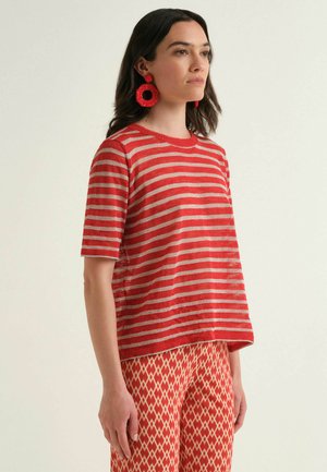 Femme aux cheveux foncés porte un haut à manches courtes rayé rouge et beige, un pantalon rouge à motif géométrique et de grandes boucles d'oreilles rouges circulaires, debout de profil.