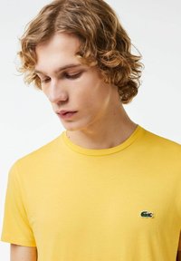 Geel katoenen T-shirt met een ronde hals en een klein groen krokodillenlogo op de linkerborst. Zachte textuur, aansluitend ontwerp.