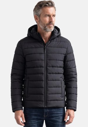 Mann mit grauem Haar und Bart, der eine schwarze gesteppte Jacke mit Kapuze und dunkelblaue Jeans trägt, steht vor einem hellen Hintergrund.