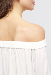 Top blanco sin mangas con escote caído y textura suave, que resalta un área de hombro y cuello liso.