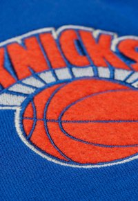 Blauer Stoff mit gesticktem orangefarbenem Basketball-Motiv und dem Text "KNICKS" in Weiß. Die Textur ist weich und weist lebendige Farben sowie klare Details auf.