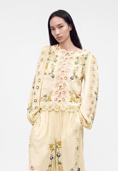 Femme aux longs cheveux foncés portant un chemisier brodé de fleurs de couleur crème et un pantalon ample assorti avec des détails en dentelle, debout devant un fond uni.
