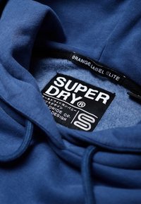 Námorně modrá mikina s kapucí a měkkým vnitřkem, s černým štítkem s bílým textem: "SUPERDRY" a slovy "ORANGE LABEL ELITE."