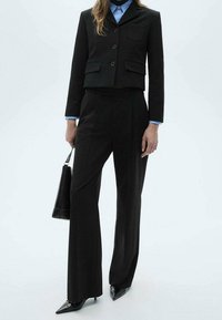 Blazer noir court avec deux poches avant, associé à un pantalon large noir. Chemise bleu clair mettant en valeur la tenue. Sac à main noir et escarpins pointus.