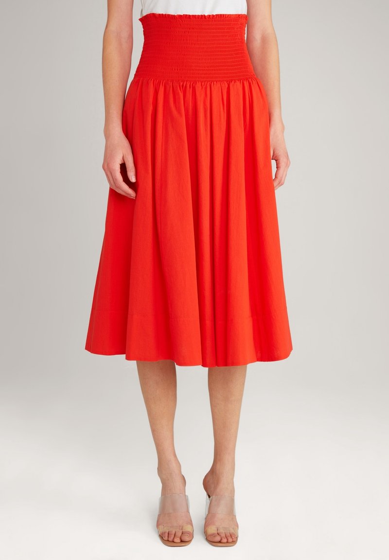 Jupe en coton rouge avec une taille haute smockée et une coupe évasée longueur midi. Portée avec des sandales à talons blocs transparents.