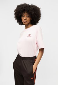 Γυναίκα με κατσαρά μαλλιά που φοράει ένα ανοιχτό ροζ t-shirt Adidas και μαύρο παντελόνι Adidas με κόκκινες ρίγες, στέκεται με τα χέρια στις τσέπες.