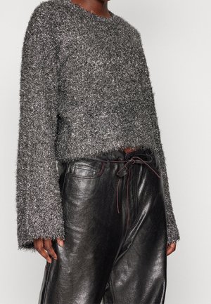 Personne portant un pull gris texturé à manches longues et un pantalon en cuir noir brillant avec une ceinture nouée à l'avant, debout devant un fond uni.