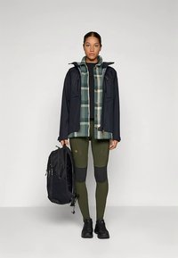 Väliväljaanne, millel on mereväe veekindel jope, roheline ruuduline särk, must ülaosa, oliivrohelised retuusid ja must seljakott. Tekstureeritud kangaste detalje.