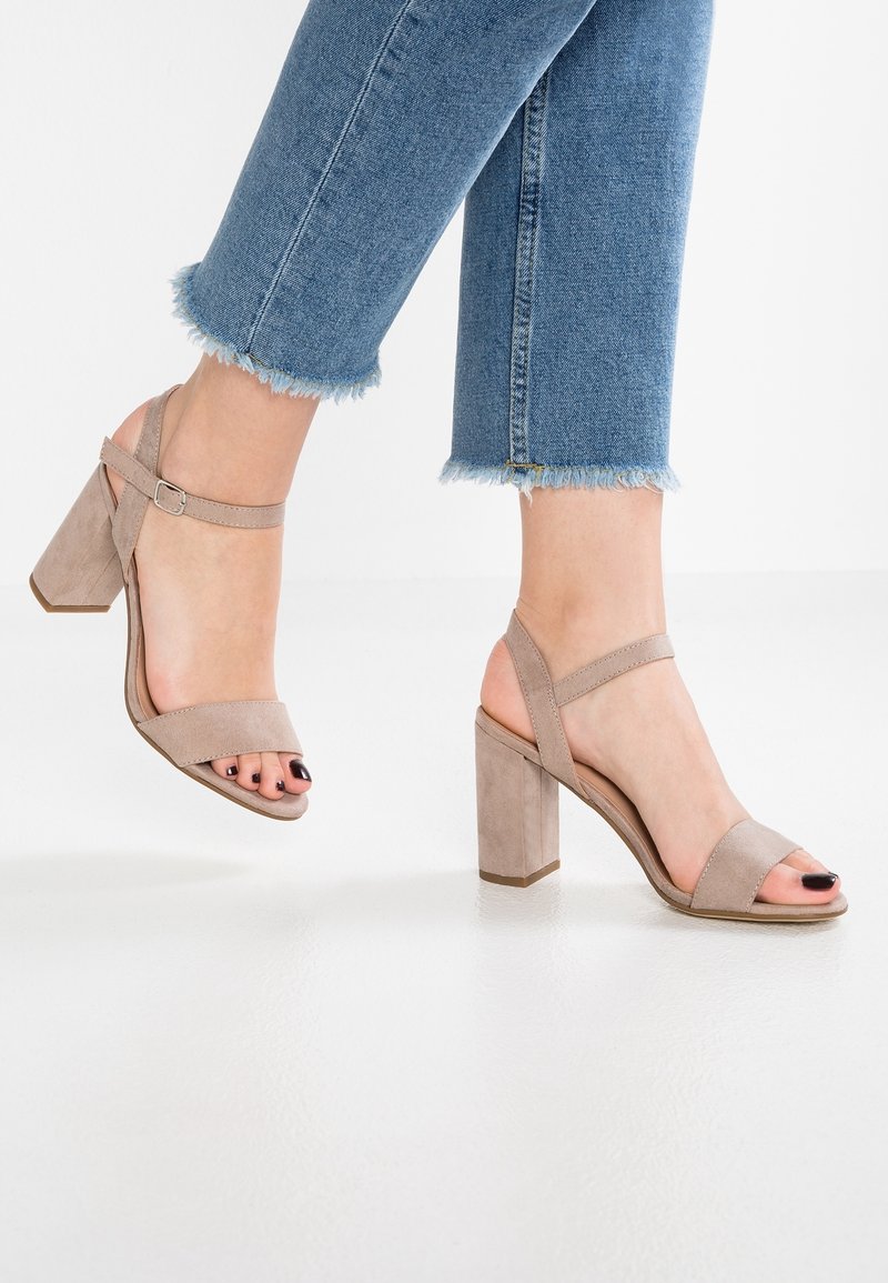 Beżowe zamszowe sandały na obcasie z szerokimi paskami z przodu i regulowanymi paskami na kostce. Blokowy obcas, prezentujący polerowaną teksturę i otwarty czubek.