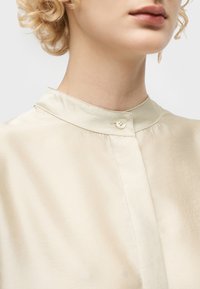 Κοντινό πλάνο ενός ατόμου που φοράει κρεμ σατέν μπλούζα με γιακά μανταρίνι που κλείνει με ένα μικρό κουμπί.