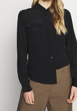 Femme portant une chemise noire à manches longues boutonnée avec des poches poitrine et un pantalon marron taille haute sur un fond uni.