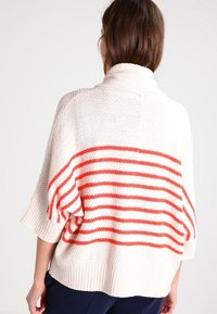 Krémový svetr s vysokým límcem, který má výrazné červené pruhy na zádech a volné rukávy, vyrobený z texturovaného pleteného materiálu.