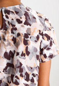 Top a maniche corte con scollatura rotonda, caratterizzato da un discreto dettaglio a chiave. Il tessuto presenta un motivo astratto tenue nei toni di grigio, marrone e beige.