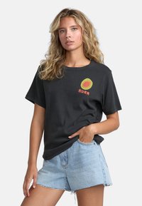 Sort bomulds t-shirt med en rund grøn og orange grafik på venstre bryst, parret med lyse denimshorts. Korte ærmer og afslappet pasform.