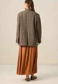 Geboekte blazer in bruin met een gestructureerde pasvorm, gecombineerd met een lange, vloeiende oranje rok en gestructureerde donkere schoenen, gezien vanaf de achterkant.