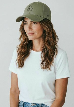 Woman with wavy brown hair wearing an olive green LA baseball cap, white t-shirt, and blue jeans, looking slightly to the right.