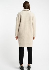 Beżowy wełniany płaszcz o fakturowanym, puszystym wzorze, z wywijanym kołnierzem i długimi rękawami, zestawiony z czarnymi wąskimi spodniami i butami.