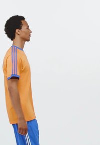 Camiseta atlética naranja con rayas azules en los hombros, combinada con pantalones cortos azules. Textura suave, mangas cortas y un corte relajado.