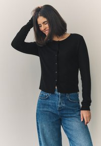 Black long-sleeve cardigan with ribbed texture and button closure, paired with light blue high-waisted jeans. Simple, fitted design.