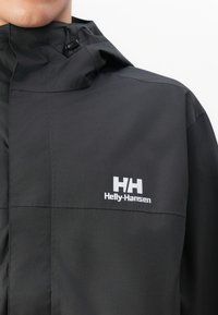 Kratak prikaz crne jakne Helly Hansen s visokim ovratnikom i bijelim logotipom na prsima, koju nosi osoba na bijeloj pozadini.