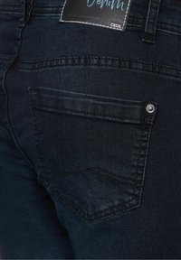 Dunkelblaue Jeans mit einer Hintertasche, die einen Metallknopf, dezente Nähte und ein Stoffetikett in der Nähe des Hosenbunds aufweist.