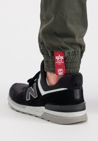 Zapatillas negras y grises con materiales de malla y gamuza, que presentan un logo blanco y una suela gris texturizada, combinadas con pantalones jogger verde oliva.