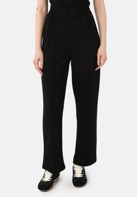 Pantalones negros de cintura alta hechos de una tela suave, con un corte relajado que presenta sutiles pliegues frontales y perneras anchas, combinados con zapatillas negras.