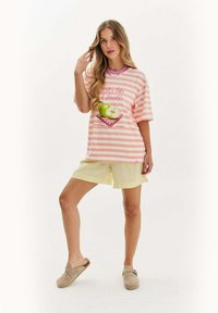 Žena stojí s jednou nohou pokrčenou, oblečená v pruhovanom tričku v ružovej a bielej farbe s grafikou zeleného jablka, svetložltých šortkách a béžových espadrilkách.
