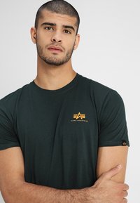 Dunkelgrünes Baumwoll-T-Shirt mit Rundhalsausschnitt, kurzen Ärmeln und einem kleinen orangefarbenen Logo auf der Brust. Verfügt über ein Markenetikett am Ärmel.