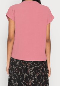 Frau in einem schlichten rosa Kurzarmoberteil und einem schwarzen Rock mit abstrakter weißer und pinker Gesichts-Linienkunst.